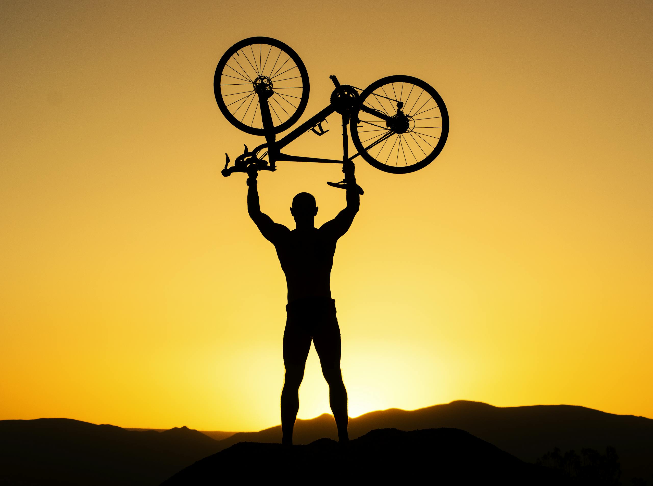 Silhouette of a strong cyclist holding a bike over their head against a stunning sunset.