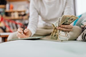 Individual counting money with pen in hand inside a cozy room setting.