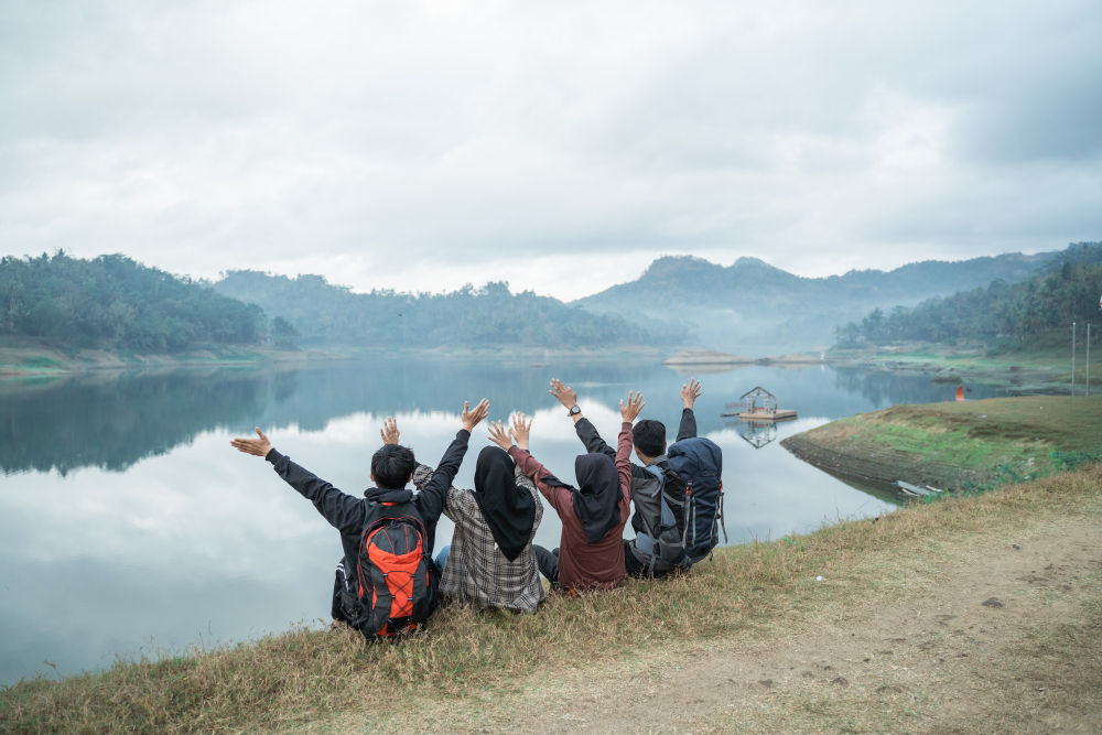 Four hikers sitting beside a lake with their arms raised.