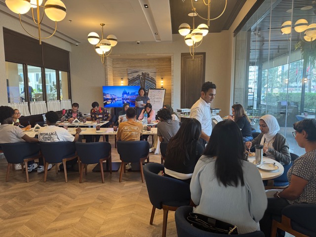 A wide room view captures students, mentors and facilitators in conversation.